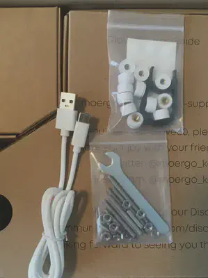 a picture of a white USB-A-to-C cable; a zip-lock bag with threads, bolts, and a mini-wrench; and a zip-lock bag with white cylinders with a screw-thread inside them, rubber feet on a sticker sheet, and a keycap puller